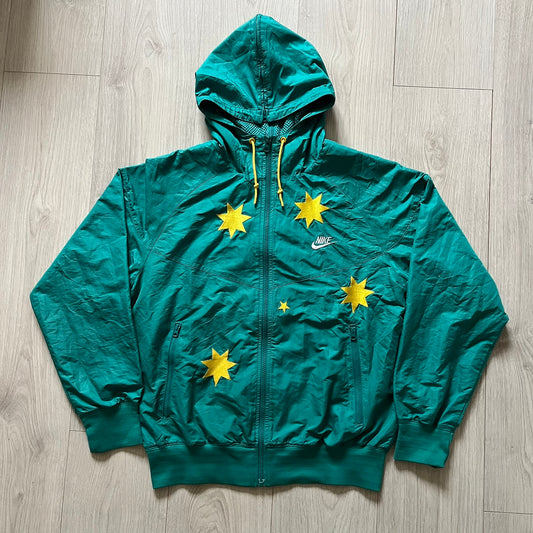 Nike 2008 Australia Olympics Split Windbreaker in teal, detailed with yellow star embroidery inspired by Australia’s national emblem. Designed with double side zippers that let the jacket separate for modular wear. Includes embroidered white Nike logo on the chest and Australian flag details inside both pockets.