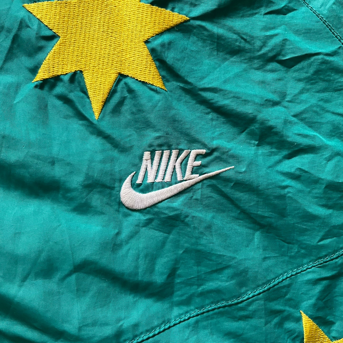 Nike 2008 Australia Olympics Split Windbreaker in teal, detailed with yellow star embroidery inspired by Australia’s national emblem. Designed with double side zippers that let the jacket separate for modular wear. Includes embroidered white Nike logo on the chest and Australian flag details inside both pockets.