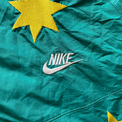 Nike 2008 Australia Olympics Split Windbreaker in teal, detailed with yellow star embroidery inspired by Australia’s national emblem. Designed with double side zippers that let the jacket separate for modular wear. Includes embroidered white Nike logo on the chest and Australian flag details inside both pockets.