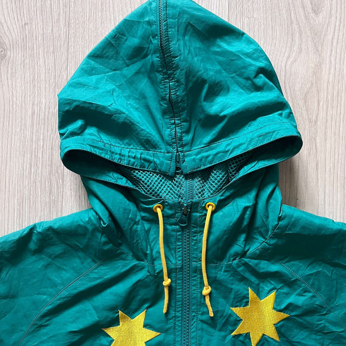 Nike 2008 Australia Olympics Split Windbreaker in teal, detailed with yellow star embroidery inspired by Australia’s national emblem. Designed with double side zippers that let the jacket separate for modular wear. Includes embroidered white Nike logo on the chest and Australian flag details inside both pockets.