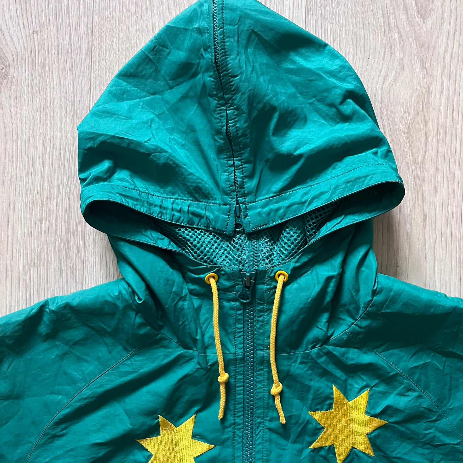 Nike 2008 Australia Olympics Split Windbreaker in teal, detailed with yellow star embroidery inspired by Australia’s national emblem. Designed with double side zippers that let the jacket separate for modular wear. Includes embroidered white Nike logo on the chest and Australian flag details inside both pockets.