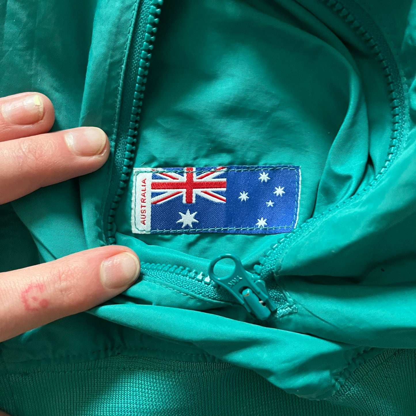Nike 2008 Australia Olympics Split Windbreaker in teal, detailed with yellow star embroidery inspired by Australia’s national emblem. Designed with double side zippers that let the jacket separate for modular wear. Includes embroidered white Nike logo on the chest and Australian flag details inside both pockets.