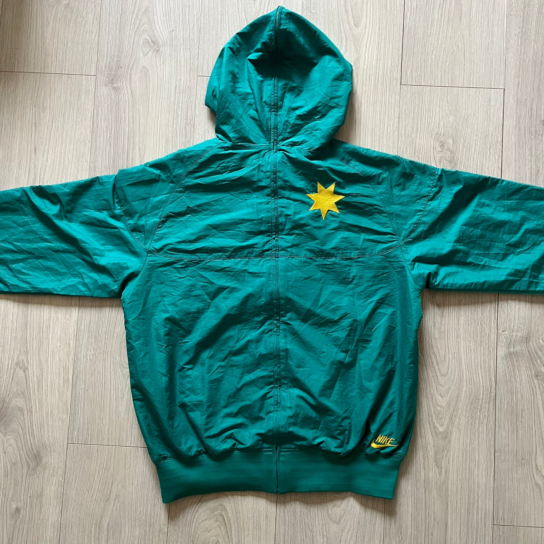 Nike 2008 Australia Olympics Split Windbreaker in teal, detailed with yellow star embroidery inspired by Australia’s national emblem. Designed with double side zippers that let the jacket separate for modular wear. Includes embroidered white Nike logo on the chest and Australian flag details inside both pockets.