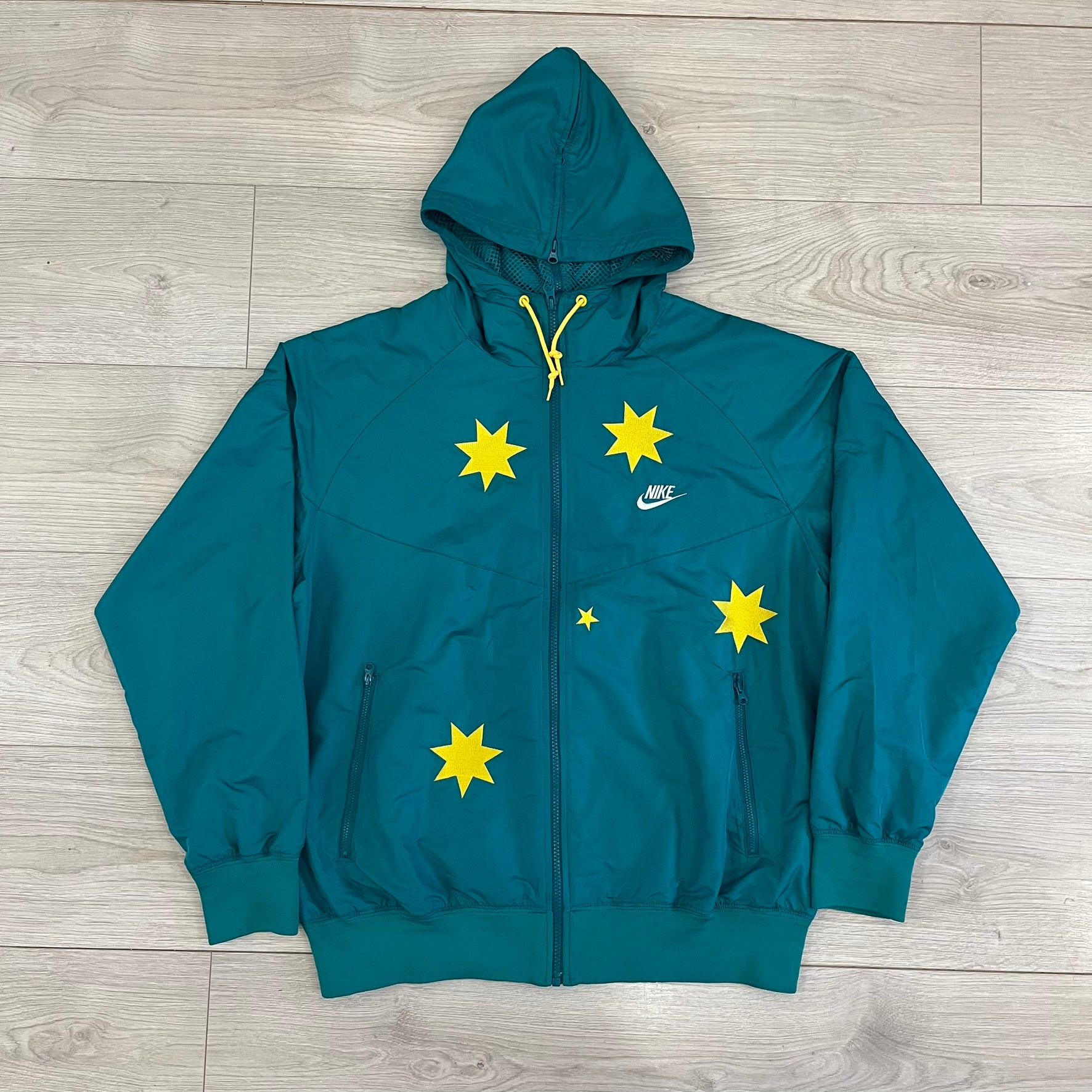 Nike 2008 Australia Olympics Split Windbreaker in teal, detailed with yellow star embroidery inspired by Australia’s national emblem. Designed with double side zippers that let the jacket separate for modular wear. Includes embroidered white Nike logo on the chest and Australian flag details inside both pockets.