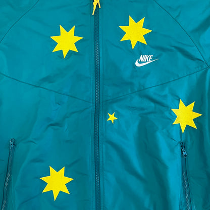 Nike 2008 Australia Olympics Split Windbreaker in teal, detailed with yellow star embroidery inspired by Australia’s national emblem. Designed with double side zippers that let the jacket separate for modular wear. Includes embroidered white Nike logo on the chest and Australian flag details inside both pockets.