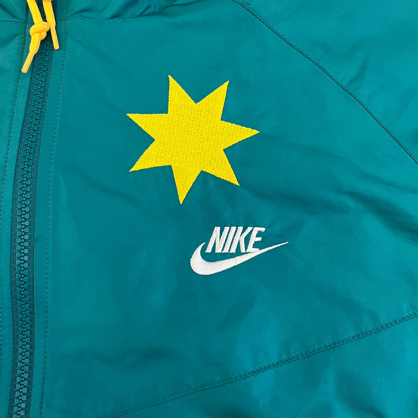 Nike 2008 Australia Olympics Split Windbreaker in teal, detailed with yellow star embroidery inspired by Australia’s national emblem. Designed with double side zippers that let the jacket separate for modular wear. Includes embroidered white Nike logo on the chest and Australian flag details inside both pockets.