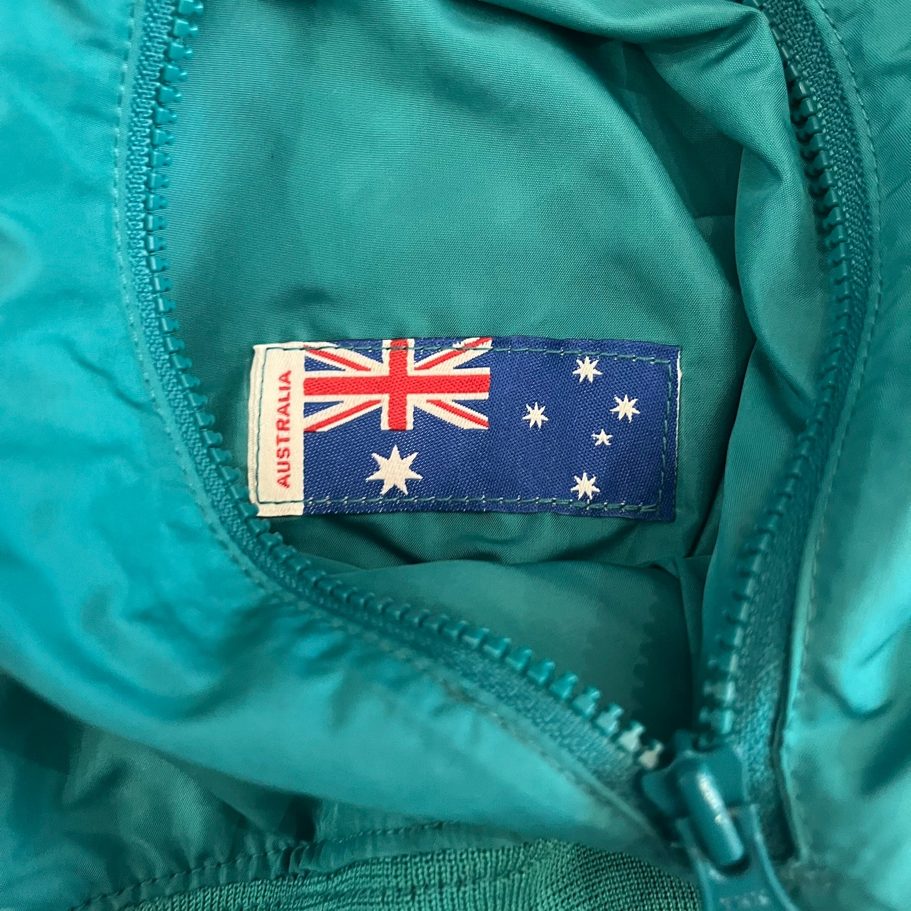 Nike 2008 Australia Olympics Split Windbreaker in teal, detailed with yellow star embroidery inspired by Australia’s national emblem. Designed with double side zippers that let the jacket separate for modular wear. Includes embroidered white Nike logo on the chest and Australian flag details inside both pockets.