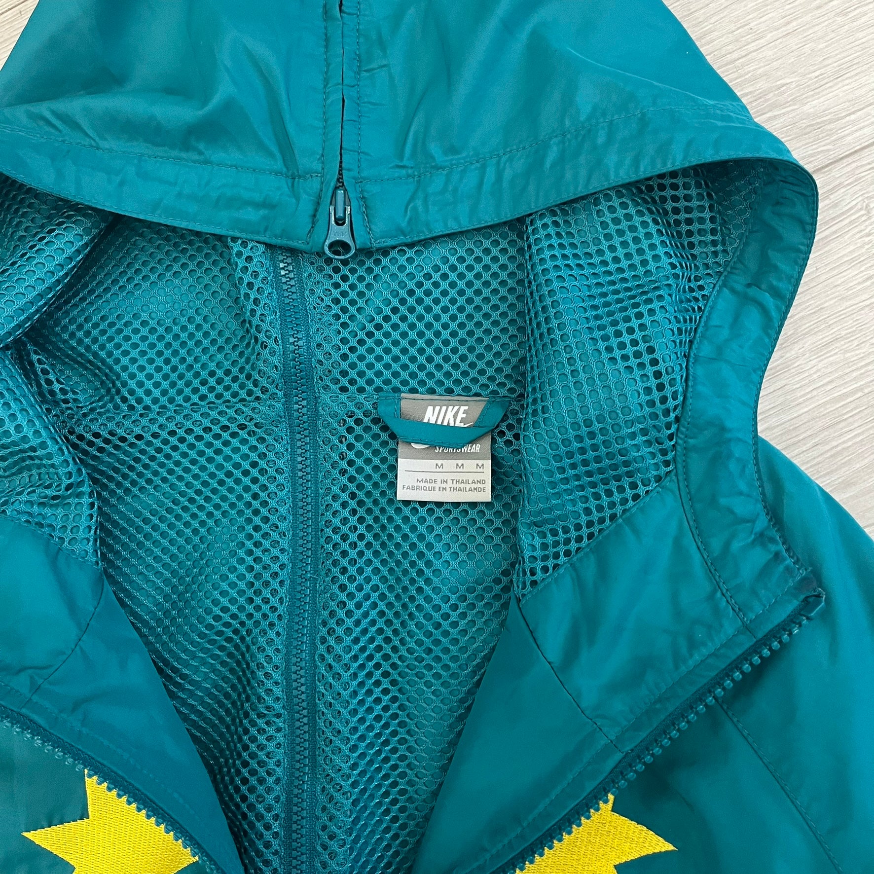 Nike 2008 Australia Olympics Split Windbreaker in teal, detailed with yellow star embroidery inspired by Australia’s national emblem. Designed with double side zippers that let the jacket separate for modular wear. Includes embroidered white Nike logo on the chest and Australian flag details inside both pockets.