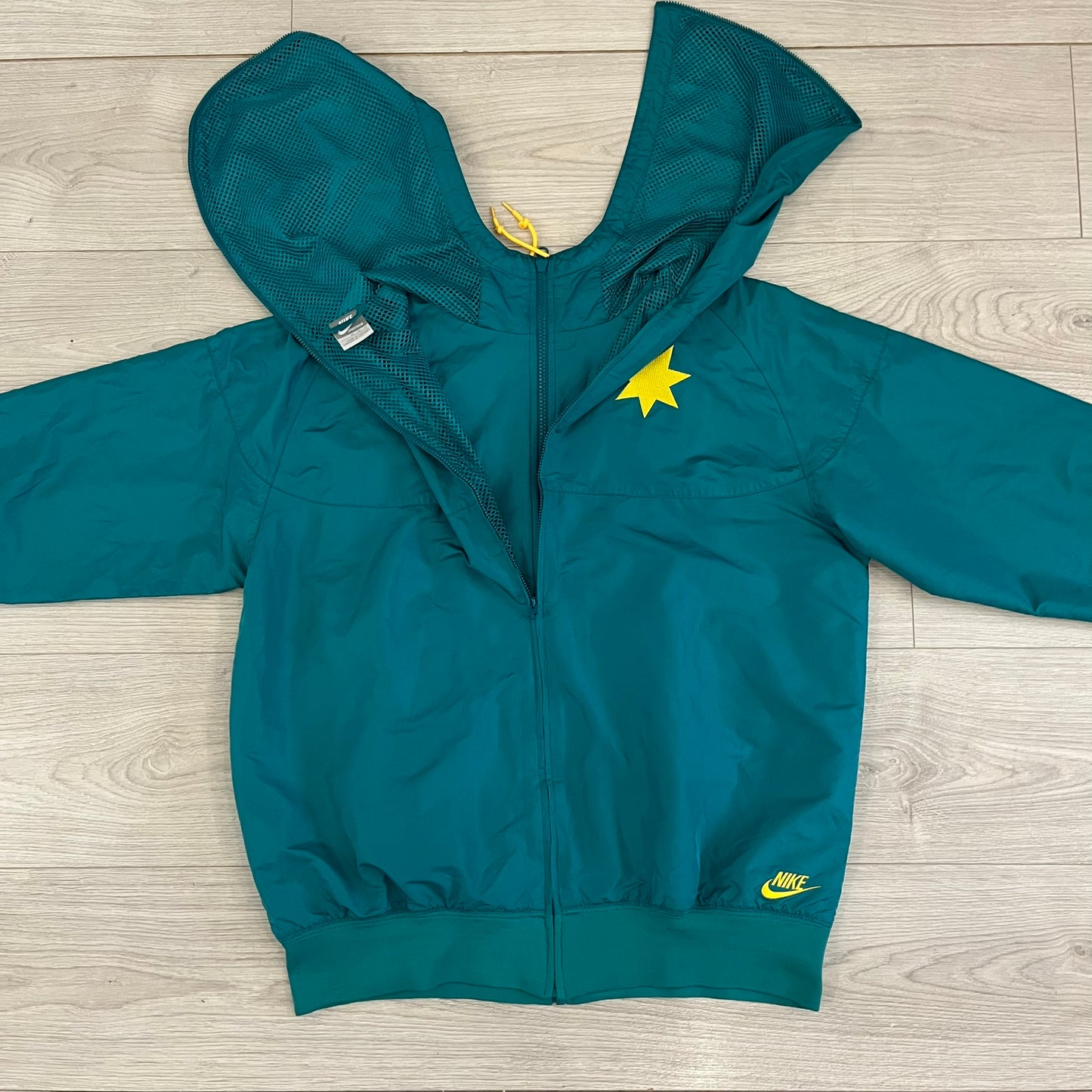 Nike 2008 Australia Olympics Split Windbreaker in teal, detailed with yellow star embroidery inspired by Australia’s national emblem. Designed with double side zippers that let the jacket separate for modular wear. Includes embroidered white Nike logo on the chest and Australian flag details inside both pockets.