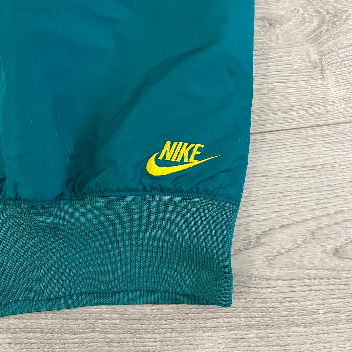 Nike 2008 Australia Olympics Split Windbreaker in teal, detailed with yellow star embroidery inspired by Australia’s national emblem. Designed with double side zippers that let the jacket separate for modular wear. Includes embroidered white Nike logo on the chest and Australian flag details inside both pockets.