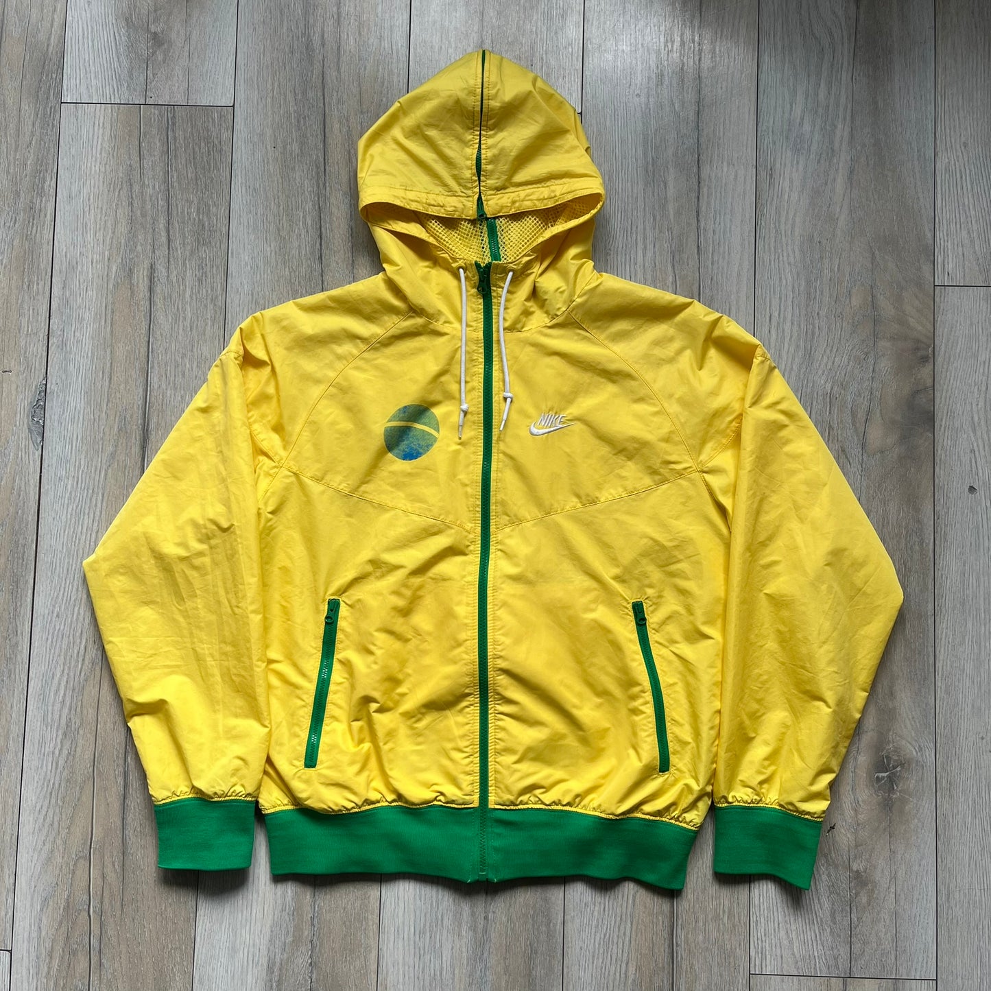 Nike Brazil Olympics Split Windbreaker from 2008, created by designer Jarrett Reynolds. Crafted in yellow with green hems and zippers. Blue circle and white Nike embroidered logo on the chest. This innovative jacket can be zipped apart on both sides and merged with other compatible models. Hidden inside, a small Brazil flags accent completes the concept.