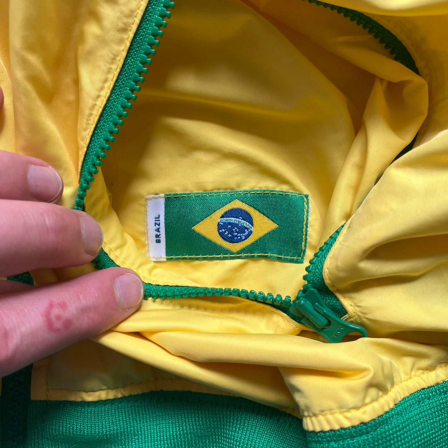 Nike Brazil Olympics Split Windbreaker from 2008, created by designer Jarrett Reynolds. Crafted in yellow with green hems and zippers. Blue circle and white Nike embroidered logo on the chest. This innovative jacket can be zipped apart on both sides and merged with other compatible models. Hidden inside, a small Brazil flags accent completes the concept.