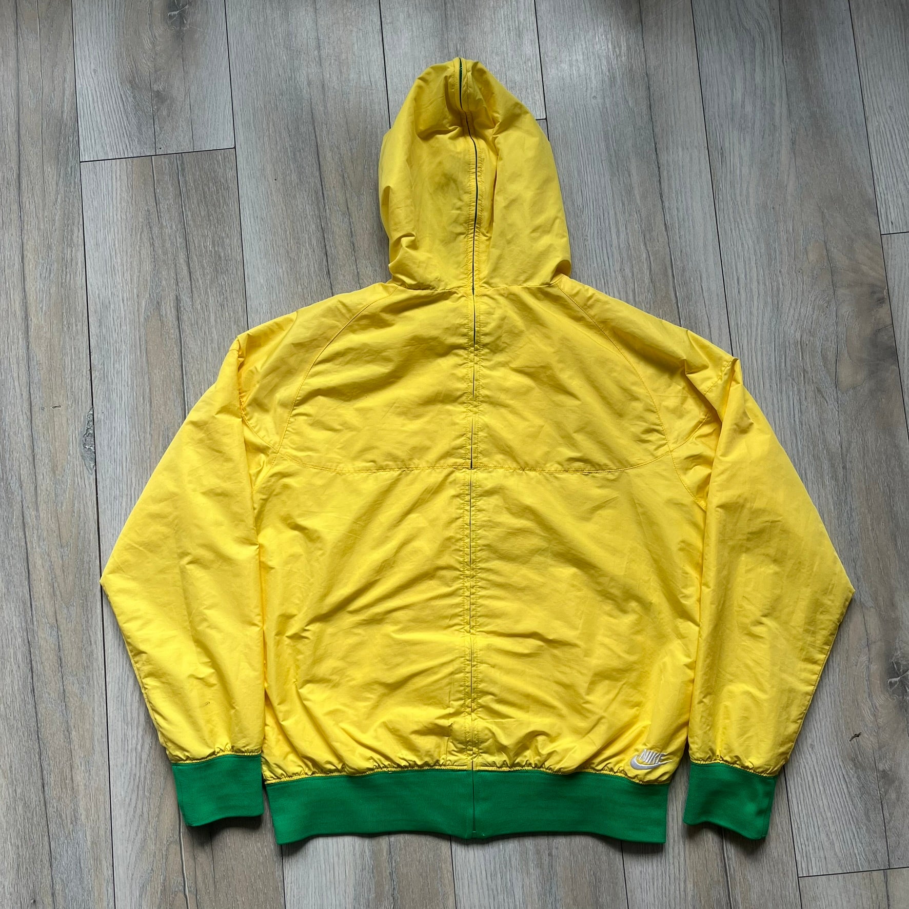 Nike Brazil Olympics Split Windbreaker from 2008, created by designer Jarrett Reynolds. Crafted in yellow with green hems and zippers. Blue circle and white Nike embroidered logo on the chest. This innovative jacket can be zipped apart on both sides and merged with other compatible models. Hidden inside, a small Brazil flags accent completes the concept.