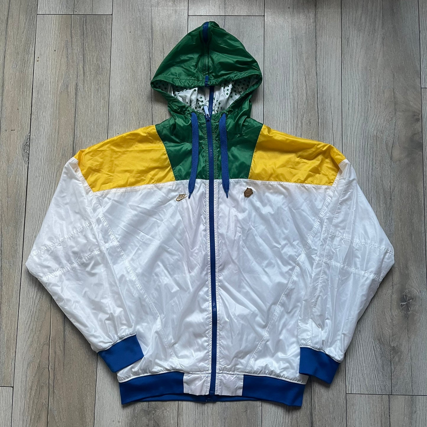 Nike Brazil windbreaker released in 2008 to mark the 50th anniversary of Brazil’s football legacy. Features a split color-block design with white lower panels and sleeves, yellow and green upper section, blue cuffs and hems, and a green hood. Detailed with a gold embroidered Nike logo on the chest, a gold-tone metal Brazil crest pin on the opposite side, and “1958-2008” with a “50” printed on the hood. The hood interior is white with an all-over green star pattern.