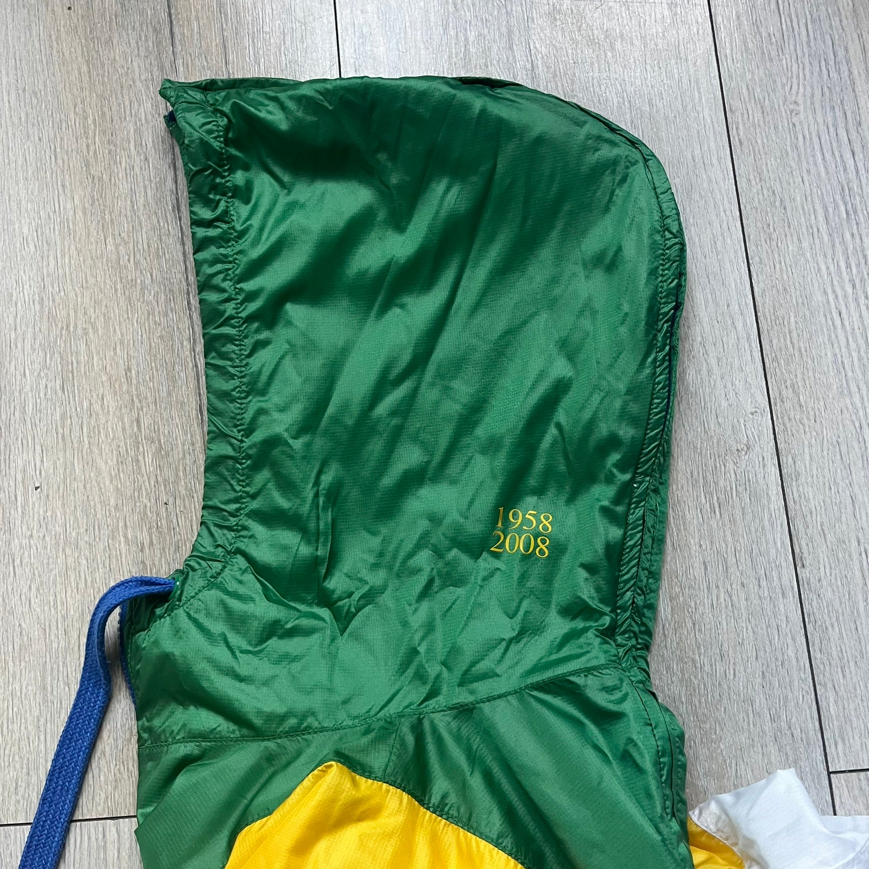 Nike Brazil windbreaker released in 2008 to mark the 50th anniversary of Brazil’s football legacy. Features a split color-block design with white lower panels and sleeves, yellow and green upper section, blue cuffs and hems, and a green hood. Detailed with a gold embroidered Nike logo on the chest, a gold-tone metal Brazil crest pin on the opposite side, and “1958-2008” with a “50” printed on the hood. The hood interior is white with an all-over green star pattern.