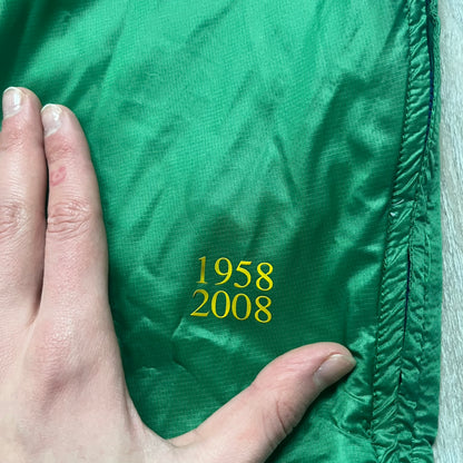 Nike Brazil windbreaker released in 2008 to mark the 50th anniversary of Brazil’s football legacy. Features a split color-block design with white lower panels and sleeves, yellow and green upper section, blue cuffs and hems, and a green hood. Detailed with a gold embroidered Nike logo on the chest, a gold-tone metal Brazil crest pin on the opposite side, and “1958-2008” with a “50” printed on the hood. The hood interior is white with an all-over green star pattern.