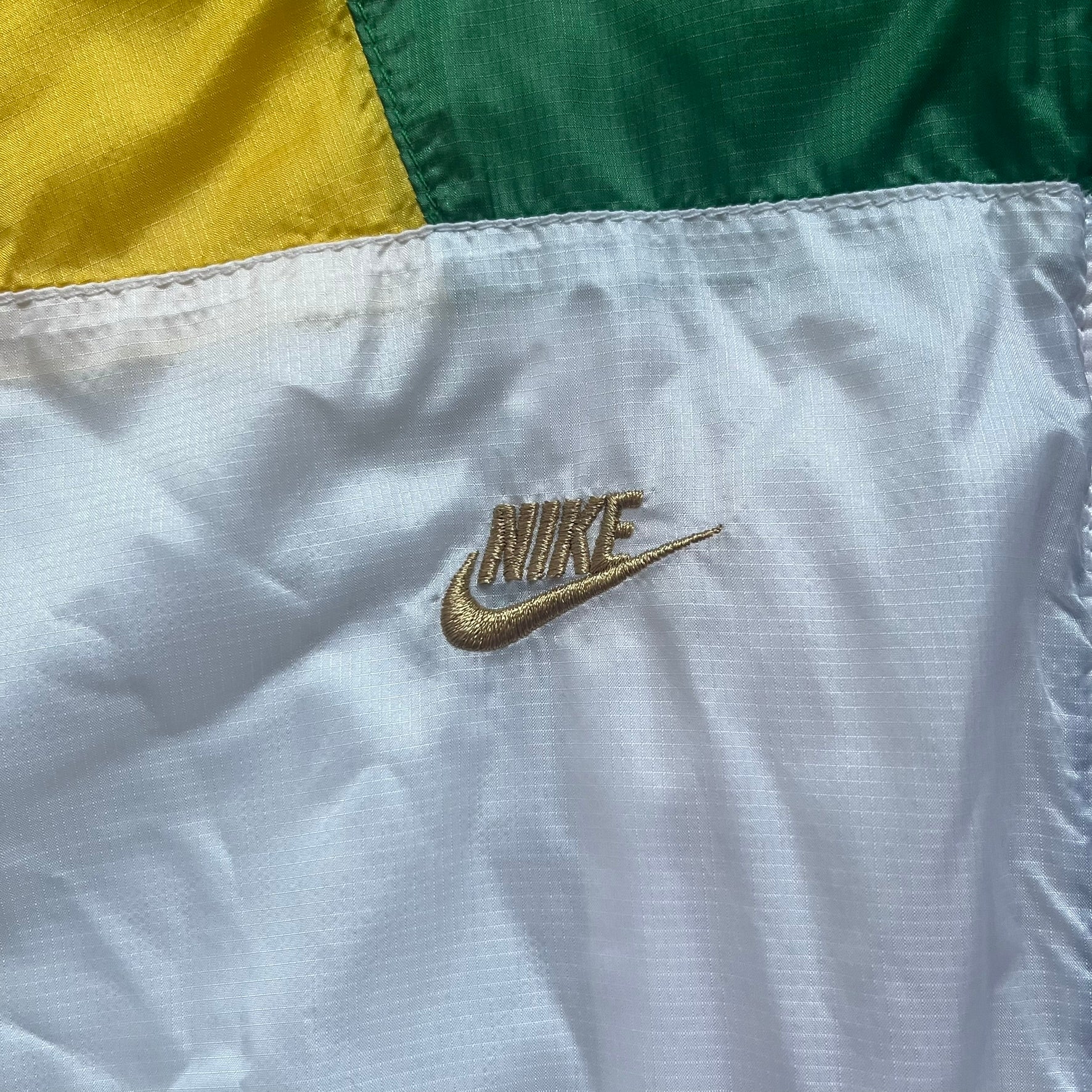 Nike Brazil windbreaker released in 2008 to mark the 50th anniversary of Brazil’s football legacy. Features a split color-block design with white lower panels and sleeves, yellow and green upper section, blue cuffs and hems, and a green hood. Detailed with a gold embroidered Nike logo on the chest, a gold-tone metal Brazil crest pin on the opposite side, and “1958-2008” with a “50” printed on the hood. The hood interior is white with an all-over green star pattern.