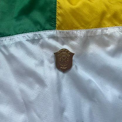 Nike Brazil windbreaker released in 2008 to mark the 50th anniversary of Brazil’s football legacy. Features a split color-block design with white lower panels and sleeves, yellow and green upper section, blue cuffs and hems, and a green hood. Detailed with a gold embroidered Nike logo on the chest, a gold-tone metal Brazil crest pin on the opposite side, and “1958-2008” with a “50” printed on the hood. The hood interior is white with an all-over green star pattern.