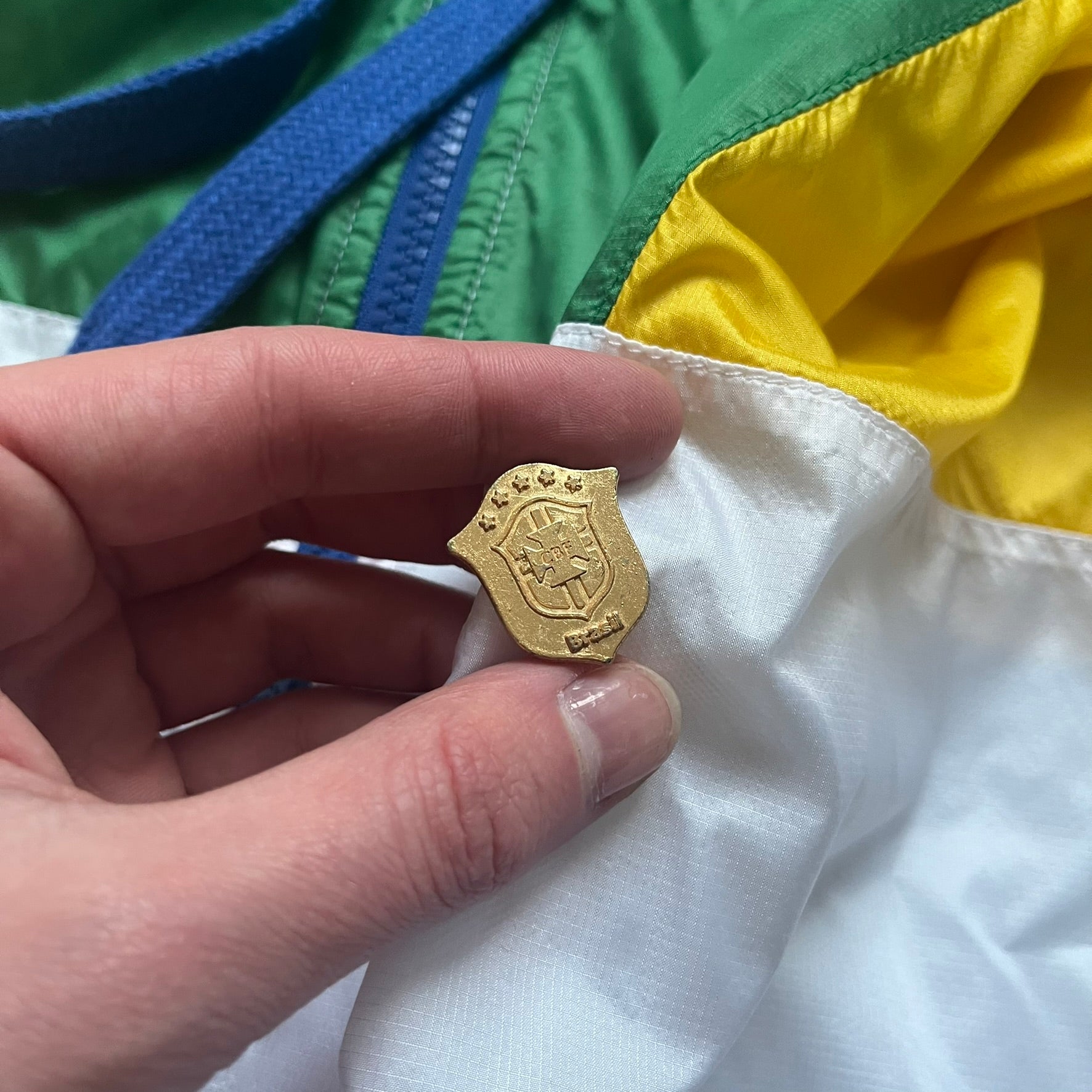 Nike Brazil windbreaker released in 2008 to mark the 50th anniversary of Brazil’s football legacy. Features a split color-block design with white lower panels and sleeves, yellow and green upper section, blue cuffs and hems, and a green hood. Detailed with a gold embroidered Nike logo on the chest, a gold-tone metal Brazil crest pin on the opposite side, and “1958-2008” with a “50” printed on the hood. The hood interior is white with an all-over green star pattern.