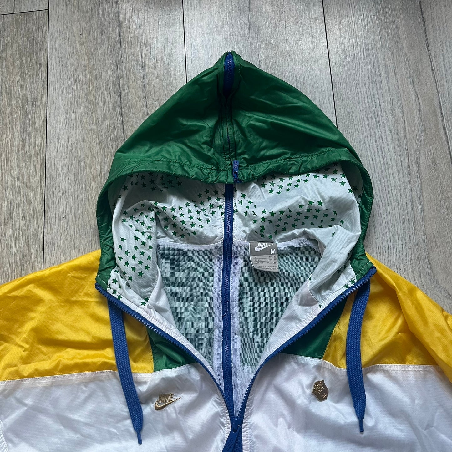 Nike Brazil windbreaker released in 2008 to mark the 50th anniversary of Brazil’s football legacy. Features a split color-block design with white lower panels and sleeves, yellow and green upper section, blue cuffs and hems, and a green hood. Detailed with a gold embroidered Nike logo on the chest, a gold-tone metal Brazil crest pin on the opposite side, and “1958-2008” with a “50” printed on the hood. The hood interior is white with an all-over green star pattern.
