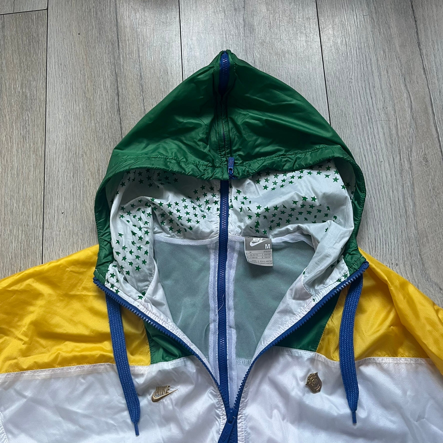 Nike Brazil windbreaker released in 2008 to mark the 50th anniversary of Brazil’s football legacy. Features a split color-block design with white lower panels and sleeves, yellow and green upper section, blue cuffs and hems, and a green hood. Detailed with a gold embroidered Nike logo on the chest, a gold-tone metal Brazil crest pin on the opposite side, and “1958-2008” with a “50” printed on the hood. The hood interior is white with an all-over green star pattern.