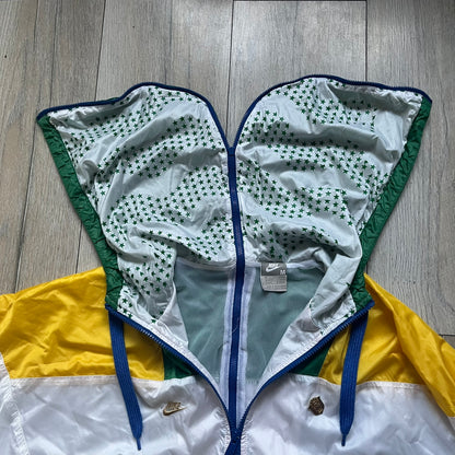Nike Brazil windbreaker released in 2008 to mark the 50th anniversary of Brazil’s football legacy. Features a split color-block design with white lower panels and sleeves, yellow and green upper section, blue cuffs and hems, and a green hood. Detailed with a gold embroidered Nike logo on the chest, a gold-tone metal Brazil crest pin on the opposite side, and “1958-2008” with a “50” printed on the hood. The hood interior is white with an all-over green star pattern.