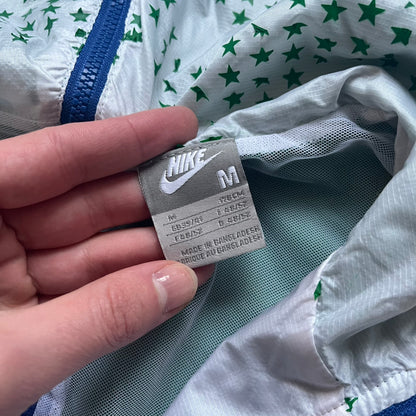 Nike Brazil windbreaker released in 2008 to mark the 50th anniversary of Brazil’s football legacy. Features a split color-block design with white lower panels and sleeves, yellow and green upper section, blue cuffs and hems, and a green hood. Detailed with a gold embroidered Nike logo on the chest, a gold-tone metal Brazil crest pin on the opposite side, and “1958-2008” with a “50” printed on the hood. The hood interior is white with an all-over green star pattern.