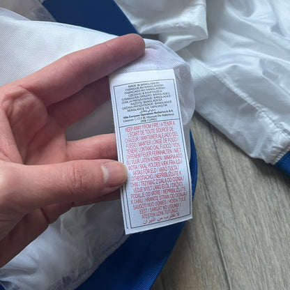 Nike Brazil windbreaker released in 2008 to mark the 50th anniversary of Brazil’s football legacy. Features a split color-block design with white lower panels and sleeves, yellow and green upper section, blue cuffs and hems, and a green hood. Detailed with a gold embroidered Nike logo on the chest, a gold-tone metal Brazil crest pin on the opposite side, and “1958-2008” with a “50” printed on the hood. The hood interior is white with an all-over green star pattern.