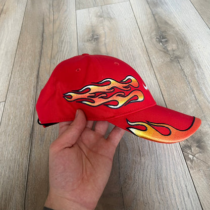 Nike Club Semi-Structured OG University Red Flame Cap is a red baseball cap featuring a white Nike Swoosh centered on the front panel. Embroidered flame graphics in orange, yellow, and red extend across the front of the visor and side panels. A small white Nike Swoosh is placed at the back. The design is identified by code IH9258-657.&nbsp;