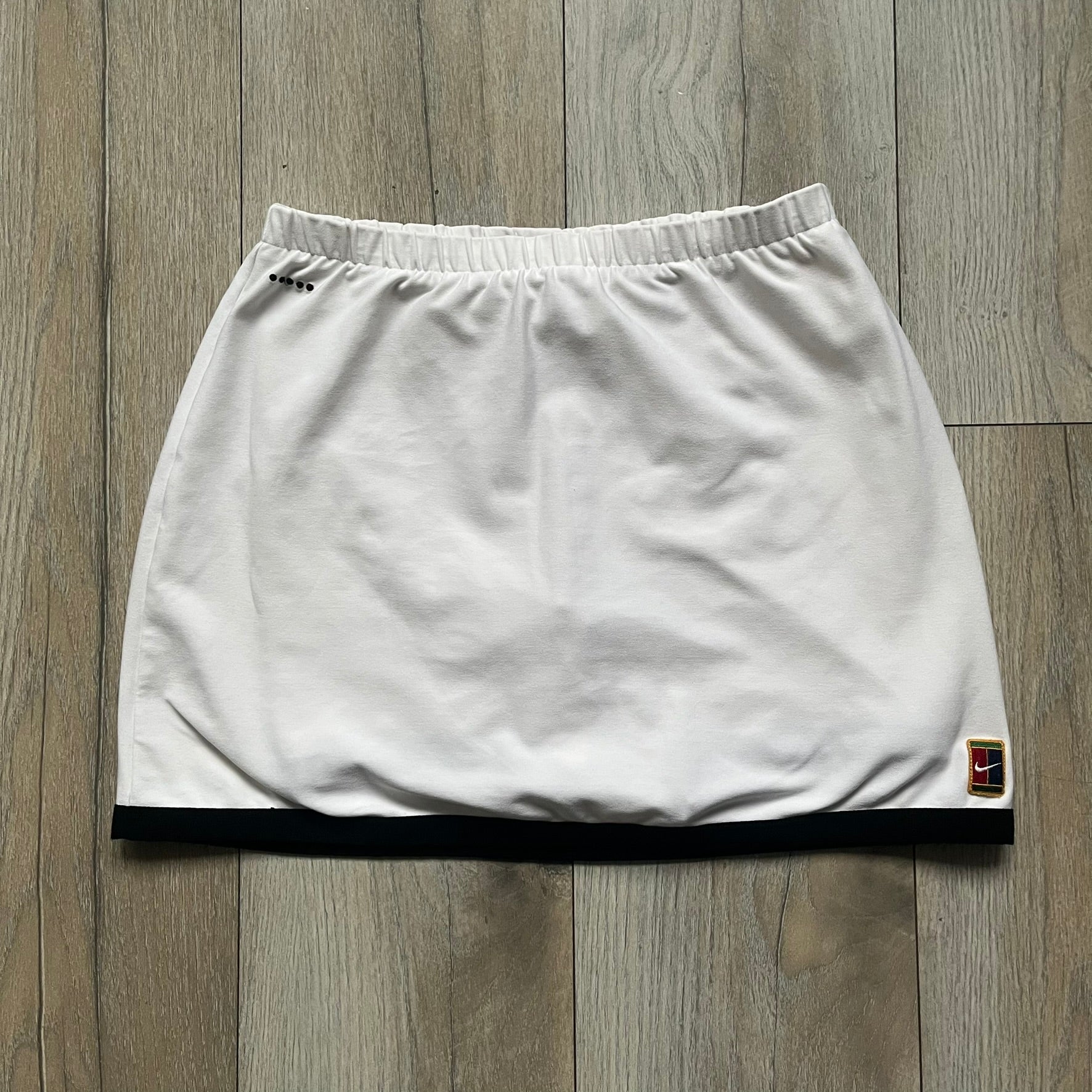 Nike Court Black Trim 90’s Tennis Skirt is a white women’s tennis skort with built-in inner shorts. It features a small Nike Court logo at the lower right hem and black trim detailing along the skirt hem and the bottom of the inner shorts legs.