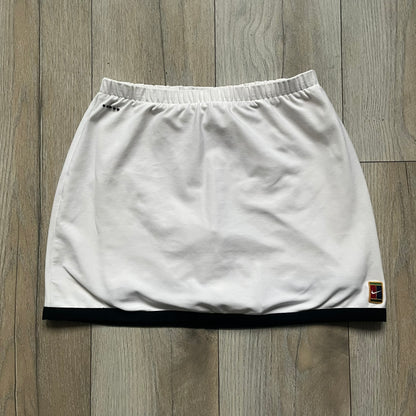 Nike Court Black Trim 90’s Tennis Skirt is a white women’s tennis skort with built-in inner shorts. It features a small Nike Court logo at the lower right hem and black trim detailing along the skirt hem and the bottom of the inner shorts legs.