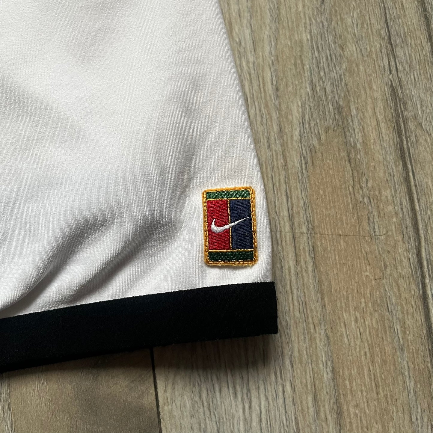 Nike Court Black Trim 90’s Tennis Skirt is a white women’s tennis skort with built-in inner shorts. It features a small Nike Court logo at the lower right hem and black trim detailing along the skirt hem and the bottom of the inner shorts legs.