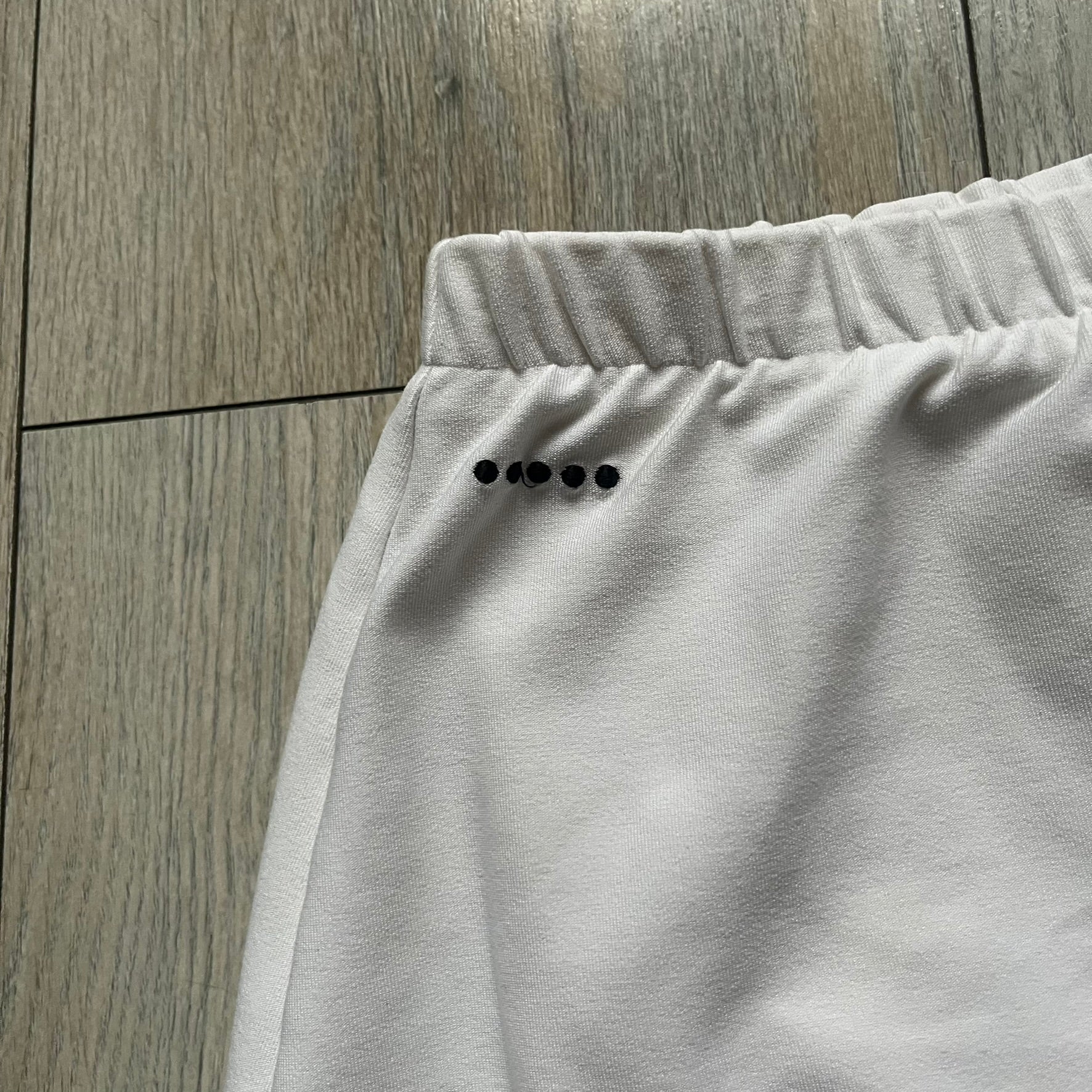 Nike Court Black Trim 90’s Tennis Skirt is a white women’s tennis skort with built-in inner shorts. It features a small Nike Court logo at the lower right hem and black trim detailing along the skirt hem and the bottom of the inner shorts legs.