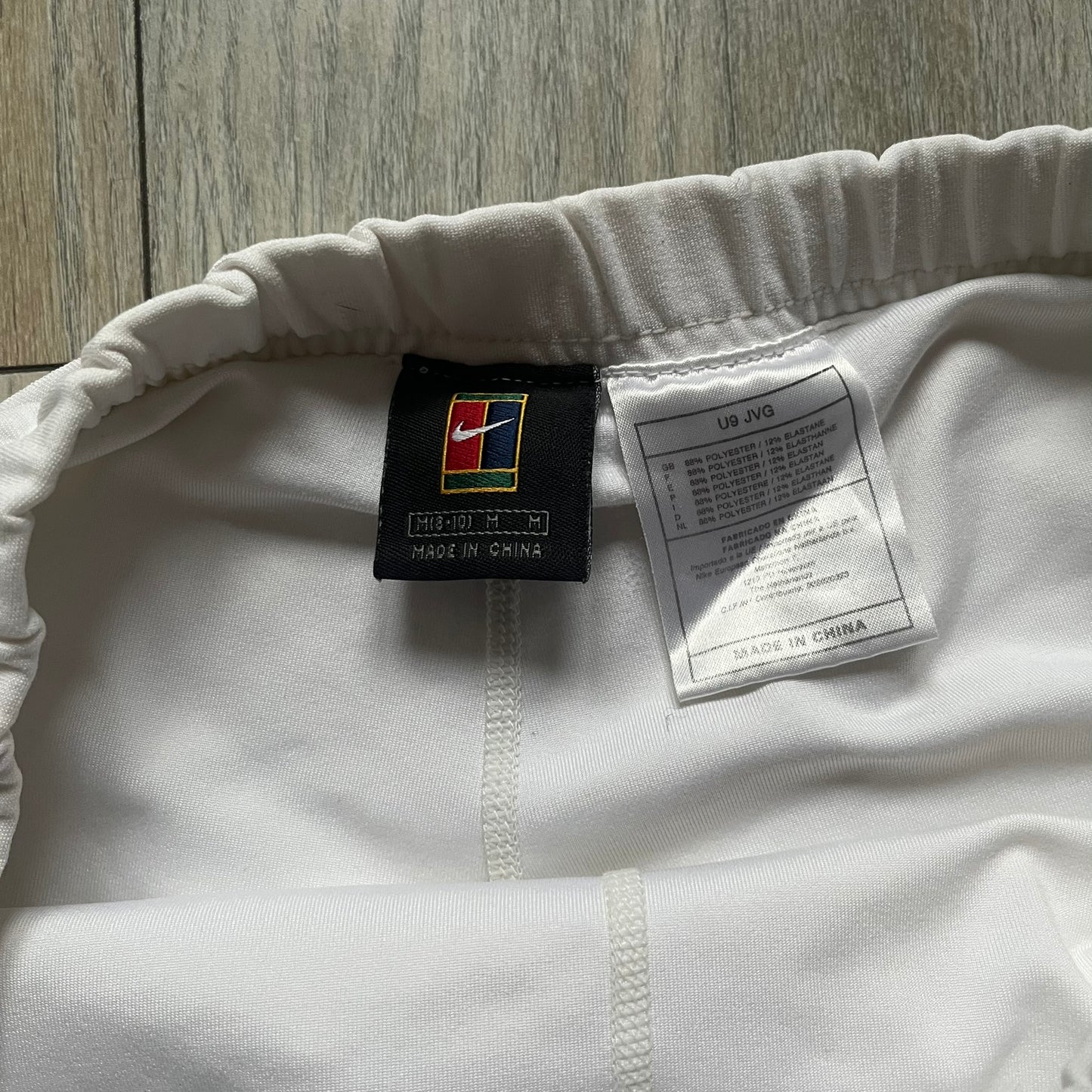 Nike Court Black Trim 90’s Tennis Skirt is a white women’s tennis skort with built-in inner shorts. It features a small Nike Court logo at the lower right hem and black trim detailing along the skirt hem and the bottom of the inner shorts legs.