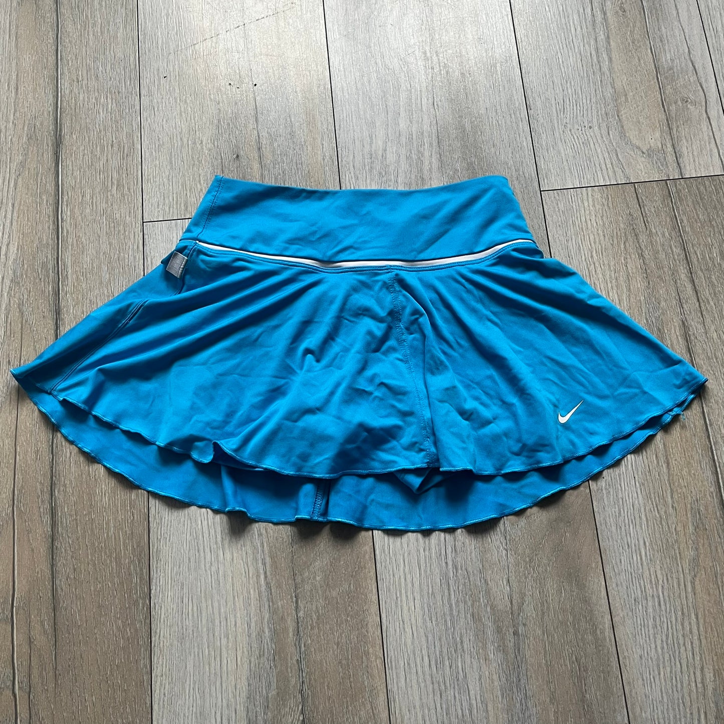 Nike Court Blue Flouncy Tennis Skirt is a women’s tennis skort featuring a built-in inner shorts layer for added coverage and support. The skirt is finished in solid blue with a small white Nike Swoosh at the lower hem and a white contrast line separating the waistband from the main body. A gray Nike Court logo is placed on the side.