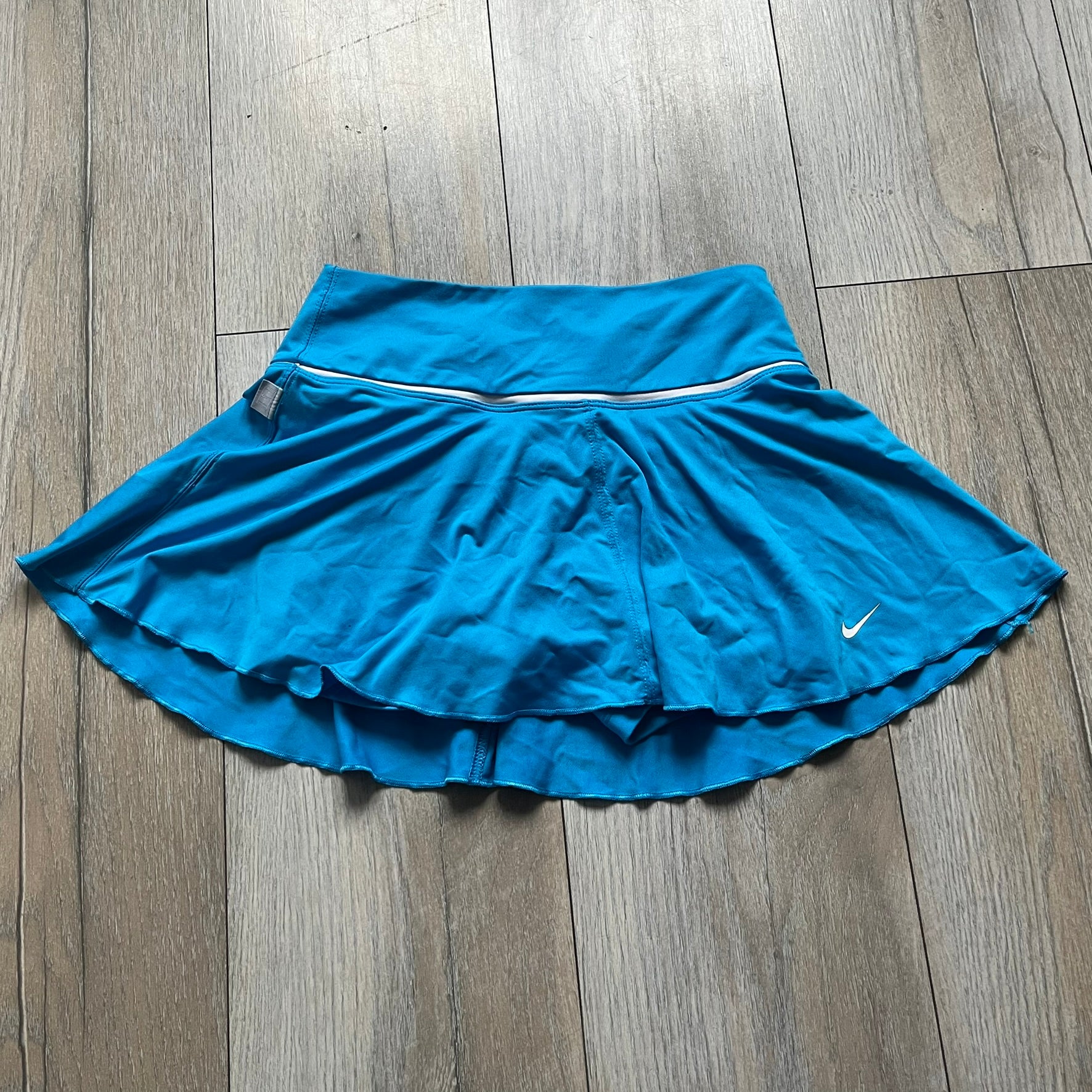 Nike Court Blue Flouncy Tennis Skirt is a women’s tennis skort featuring a built-in inner shorts layer for added coverage and support. The skirt is finished in solid blue with a small white Nike Swoosh at the lower hem and a white contrast line separating the waistband from the main body. A gray Nike Court logo is placed on the side.