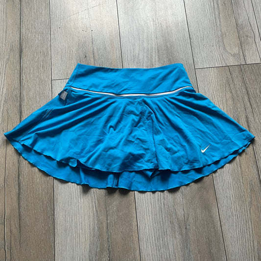 Nike Court Blue Flouncy Tennis Skirt is a women’s tennis skort featuring a built-in inner shorts layer for added coverage and support. The skirt is finished in solid blue with a small white Nike Swoosh at the lower hem and a white contrast line separating the waistband from the main body. A gray Nike Court logo is placed on the side.