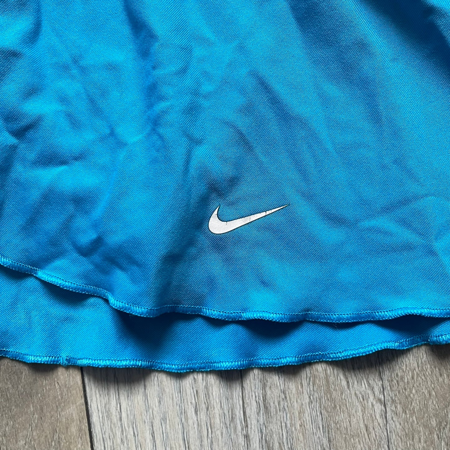 Nike Court Blue Flouncy Tennis Skirt is a women’s tennis skort featuring a built-in inner shorts layer for added coverage and support. The skirt is finished in solid blue with a small white Nike Swoosh at the lower hem and a white contrast line separating the waistband from the main body. A gray Nike Court logo is placed on the side.