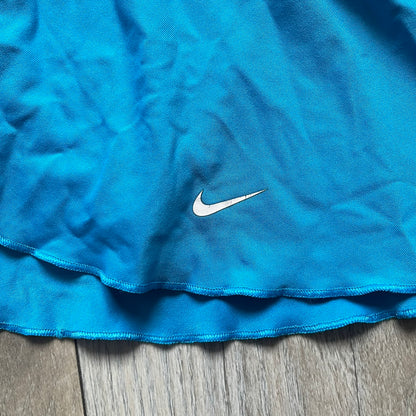 Nike Court Blue Flouncy Tennis Skirt is a women’s tennis skort featuring a built-in inner shorts layer for added coverage and support. The skirt is finished in solid blue with a small white Nike Swoosh at the lower hem and a white contrast line separating the waistband from the main body. A gray Nike Court logo is placed on the side.