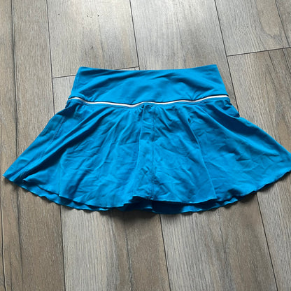 Nike Court Blue Flouncy Tennis Skirt is a women’s tennis skort featuring a built-in inner shorts layer for added coverage and support. The skirt is finished in solid blue with a small white Nike Swoosh at the lower hem and a white contrast line separating the waistband from the main body. A gray Nike Court logo is placed on the side.