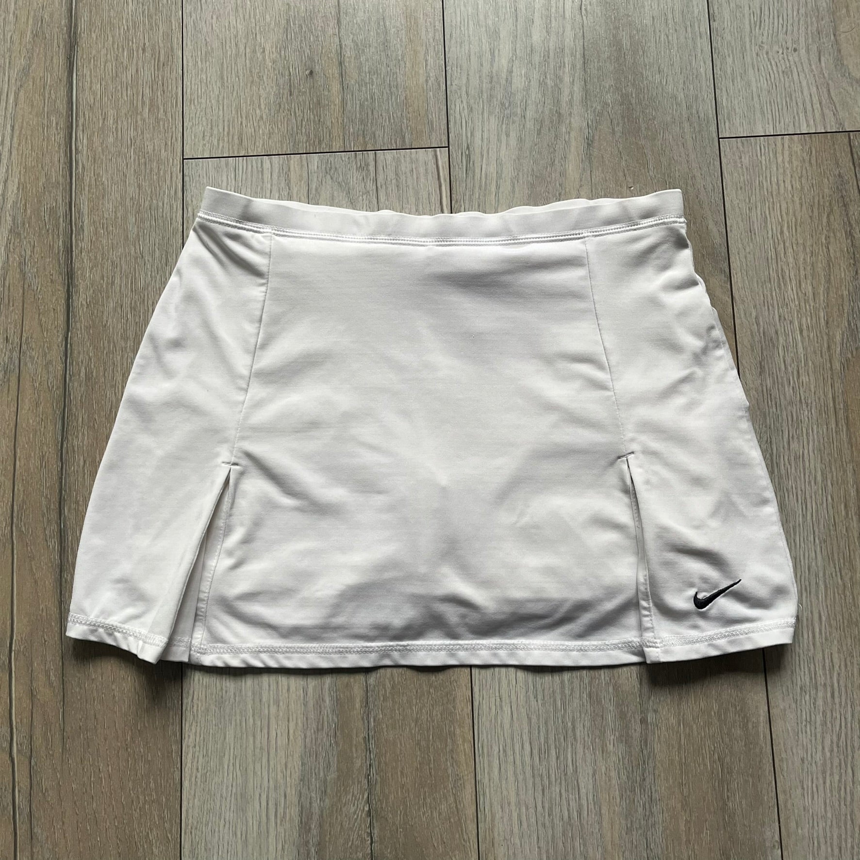 Nike Court Flex Pure Tennis Skirt is a kids’ tennis skort with integrated inner shorts for coverage and support. The skirt is white with a black Nike Swoosh at the lower hem and a Nike Court logo on the opposite side. The front features cut-out sections on both sides for added comfort and mobility.