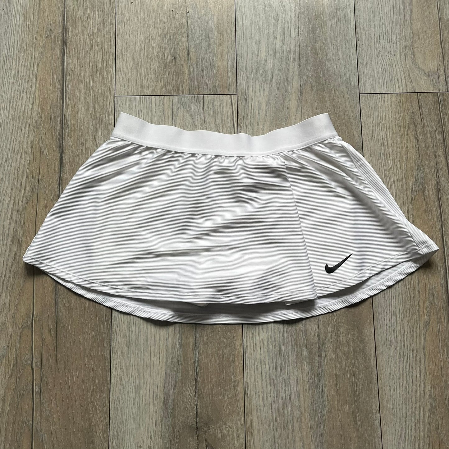 Nike Court Victory Tennis Skirt is a kids’ tennis skort with built-in inner shorts for coverage. The skirt features a textured fabric with fine linear detailing, while the waistband remains smooth, and a black Nike Swoosh is placed at the lower hem. The design includes a pleated section.