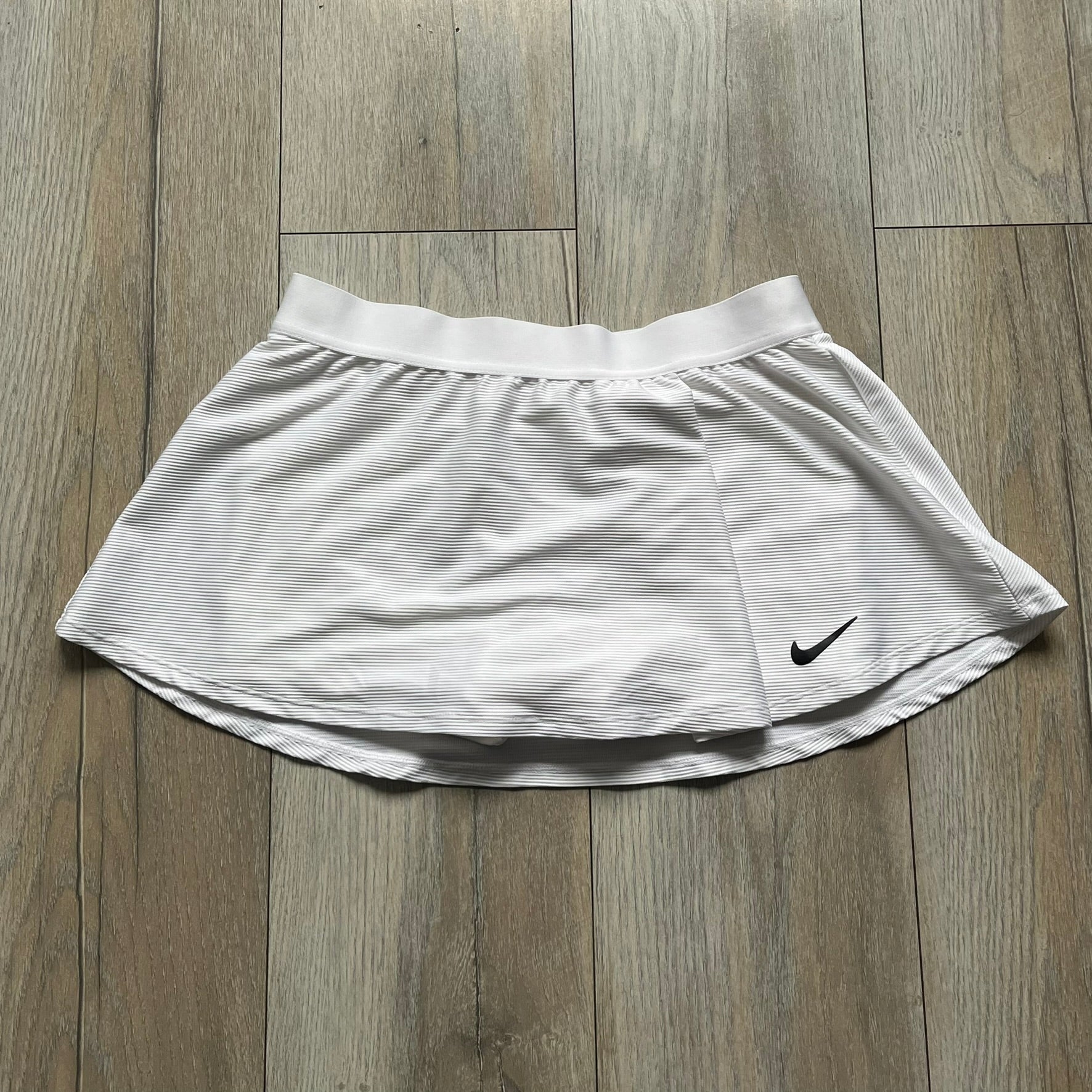 Nike Court Victory Tennis Skirt is a kids’ tennis skort with built-in inner shorts for coverage. The skirt features a textured fabric with fine linear detailing, while the waistband remains smooth, and a black Nike Swoosh is placed at the lower hem. The design includes a pleated section.