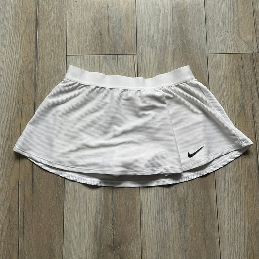 Nike Court Victory Tennis Skirt is a kids’ tennis skort with built-in inner shorts for coverage. The skirt features a textured fabric with fine linear detailing, while the waistband remains smooth, and a black Nike Swoosh is placed at the lower hem. The design includes a pleated section.