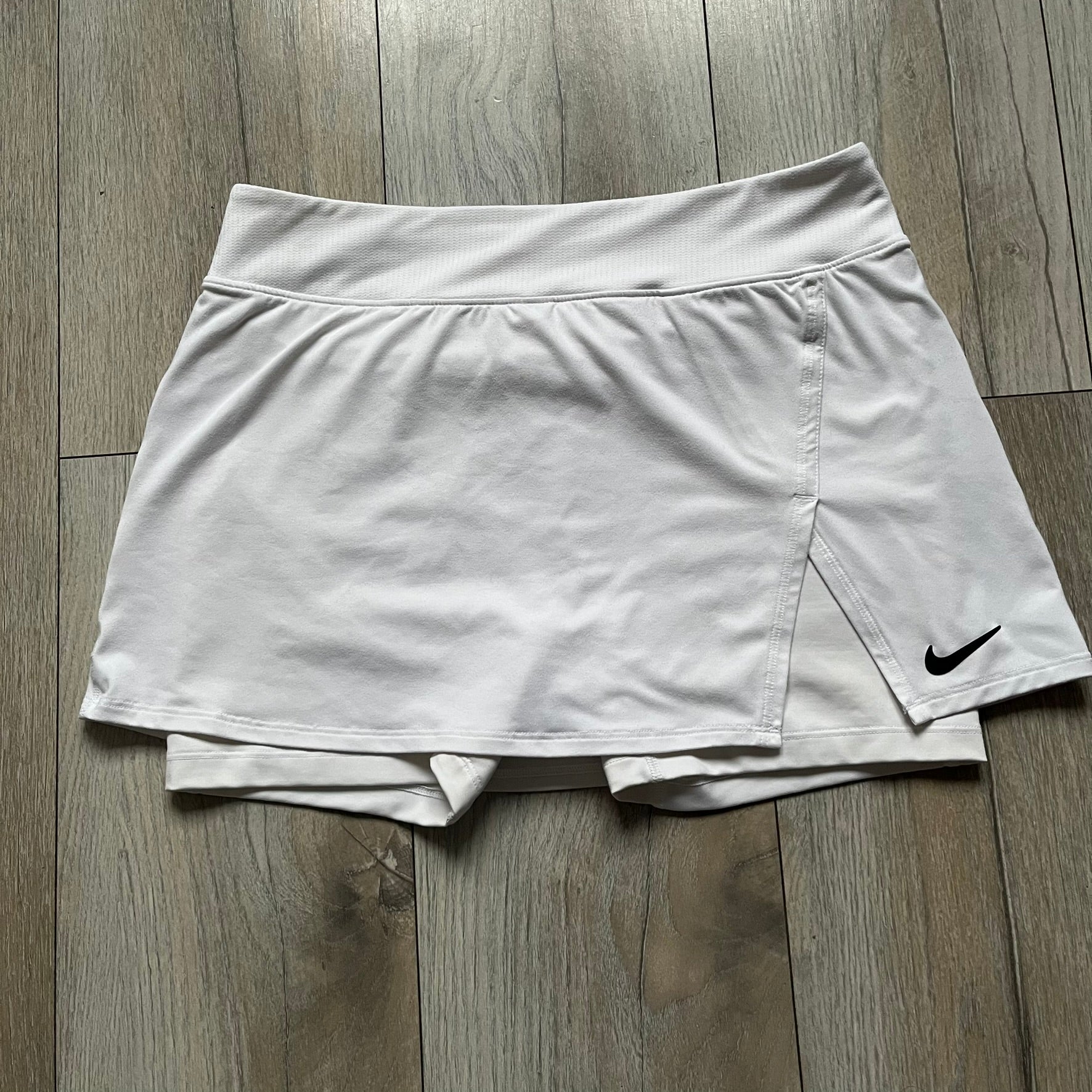 Nike Court Victory W Tennis Skirt is a women’s tennis skort with built-in inner shorts for coverage. The skirt is white with a Nike Swoosh at the lower hem and features a triangular cutout detail next to the logo for added mobility.