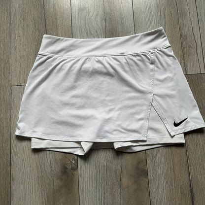 Nike Court Victory W Tennis Skirt is a women’s tennis skort with built-in inner shorts for coverage. The skirt is white with a Nike Swoosh at the lower hem and features a triangular cutout detail next to the logo for added mobility.