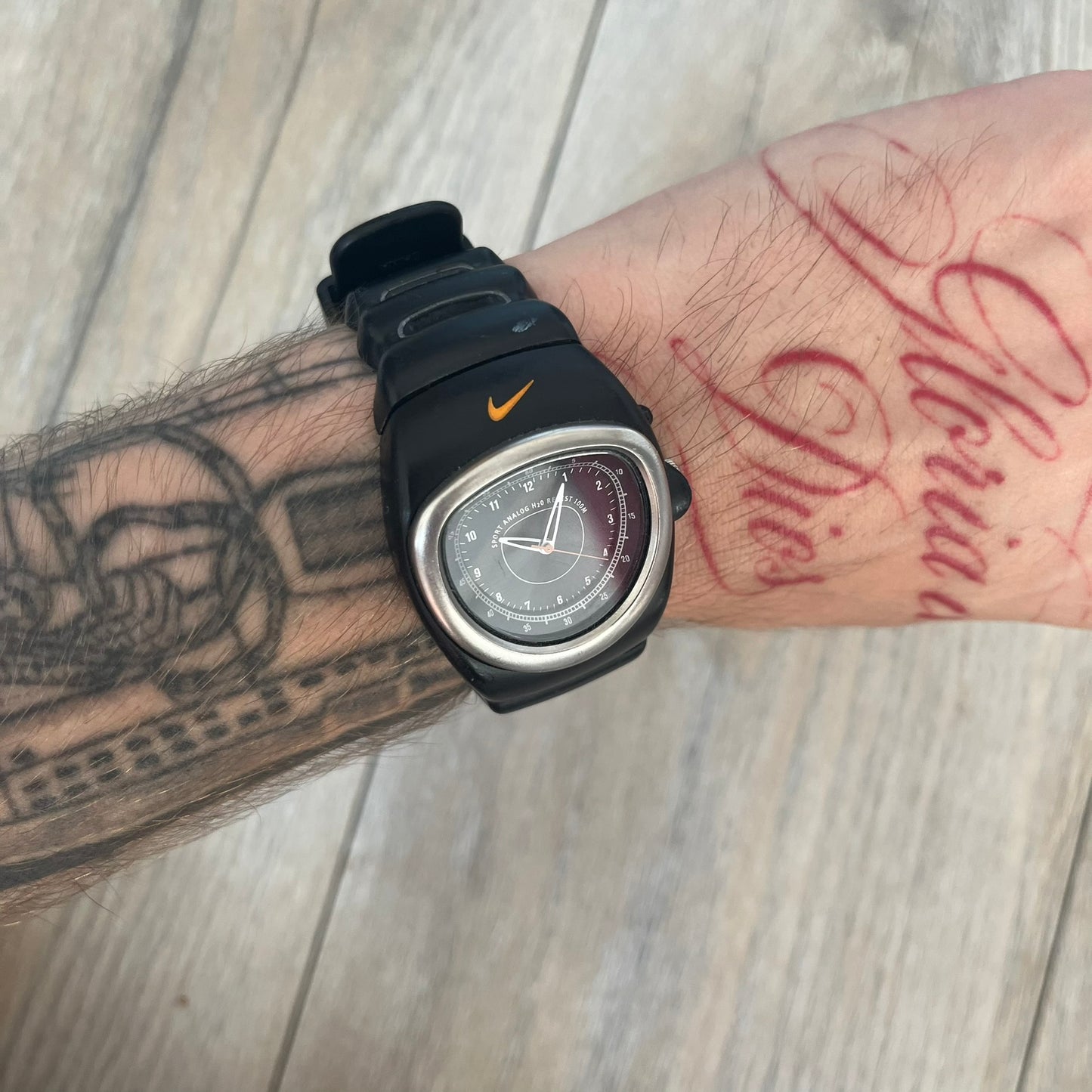 Nike Cross Training VG72-5000 Analog Watch is a 2000s model featuring a black strap, silver-tone stainless steel case, and black dial. The oval-shaped case has a flattened top with an orange Nike Swoosh positioned above the dial. It uses a three-hand analog display and includes an additional button above the crown to operate the backlight.