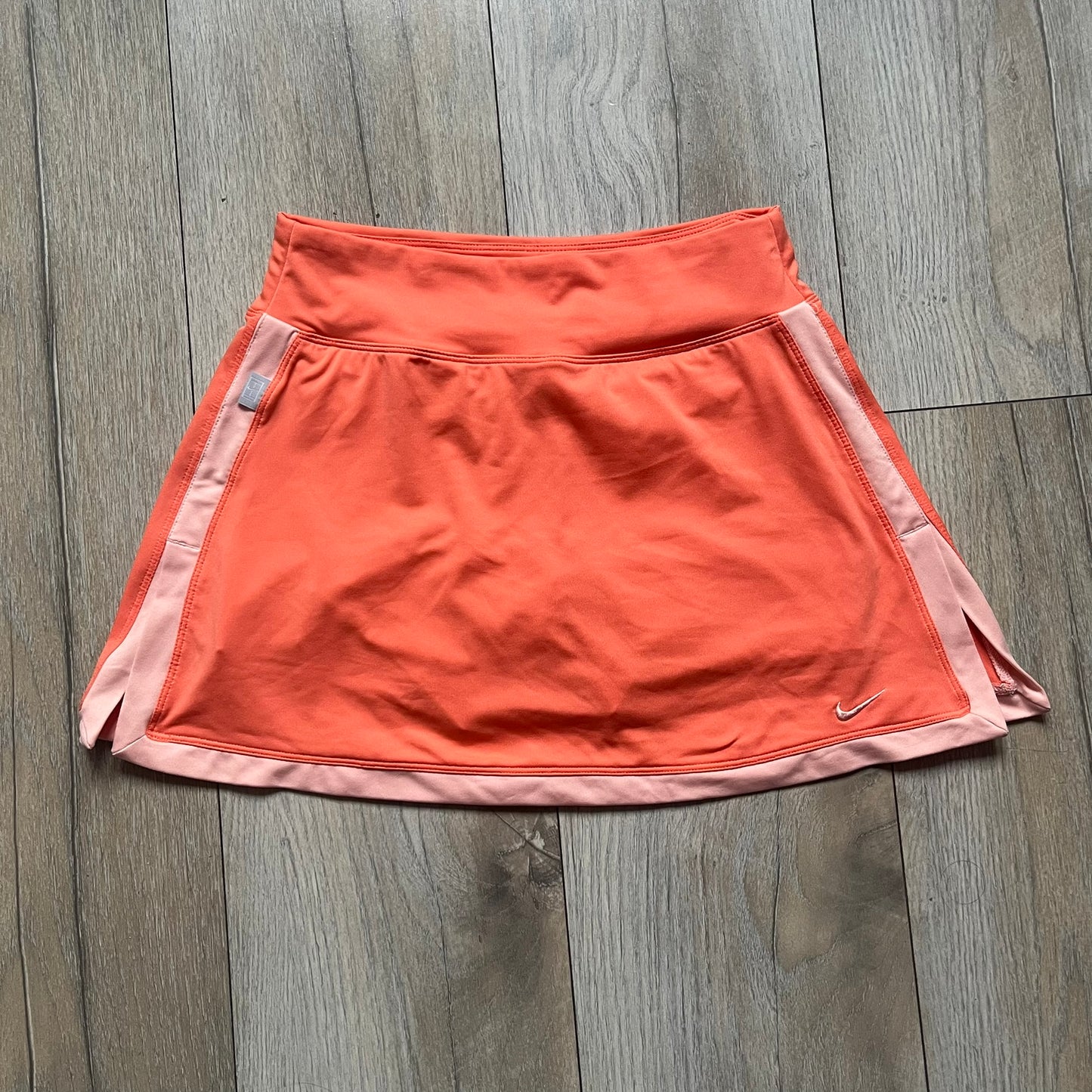 Nike Dri-FIT Orange Tennis Skirt is a women’s tennis skort with built-in shorts for coverage and support. The skirt is finished in orange with lighter side panels and embroidered Nike Swoosh at the lower hem. A small Nike Court logo on the side highlights its connection to Nike’s performance tennis line.