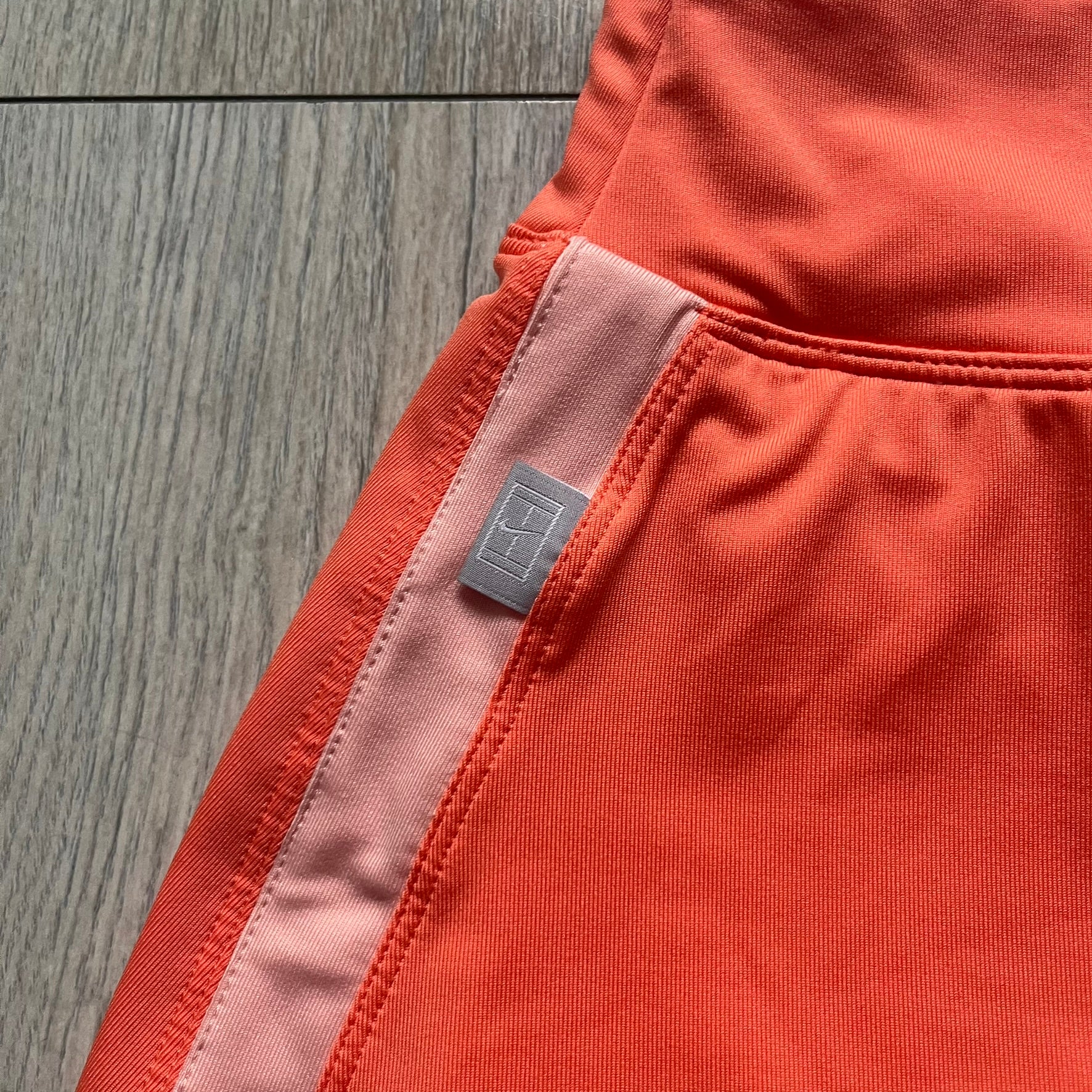 Nike Dri-FIT Orange Tennis Skirt is a women’s tennis skort with built-in shorts for coverage and support. The skirt is finished in orange with lighter side panels and embroidered Nike Swoosh at the lower hem. A small Nike Court logo on the side highlights its connection to Nike’s performance tennis line.