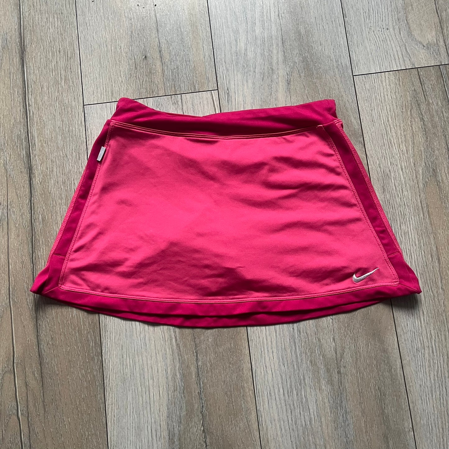 Nike Dri-FIT Pink Tennis Skirt is a women’s tennis skort with built-in shorts for coverage and support. The skirt is finished in dark pink with darker side panels and waistband, featuring a white embroidered Nike Swoosh at the lower hem. A small Nike Court logo on the side highlights its connection to Nike’s performance tennis line.